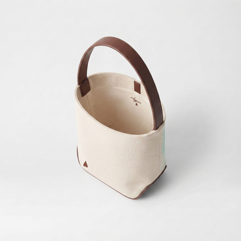 A canvas bucket bag in cream with dark brown leather trim and handle, shown from an elevated angle on a light gray background.