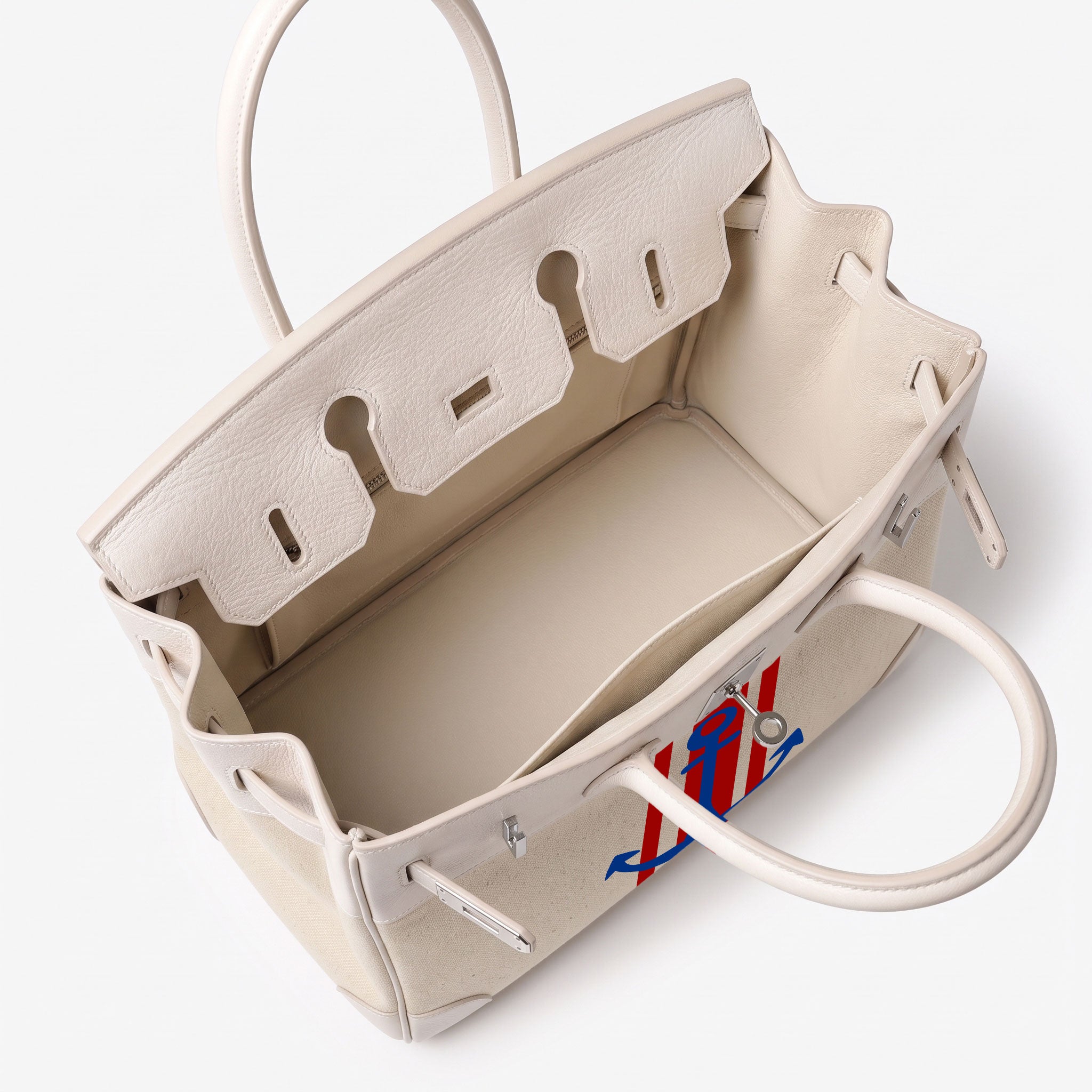 An open cream-colored leather handbag with silver-tone hardware, showcasing its interior lining and a small red and blue embroidered accent on the front.