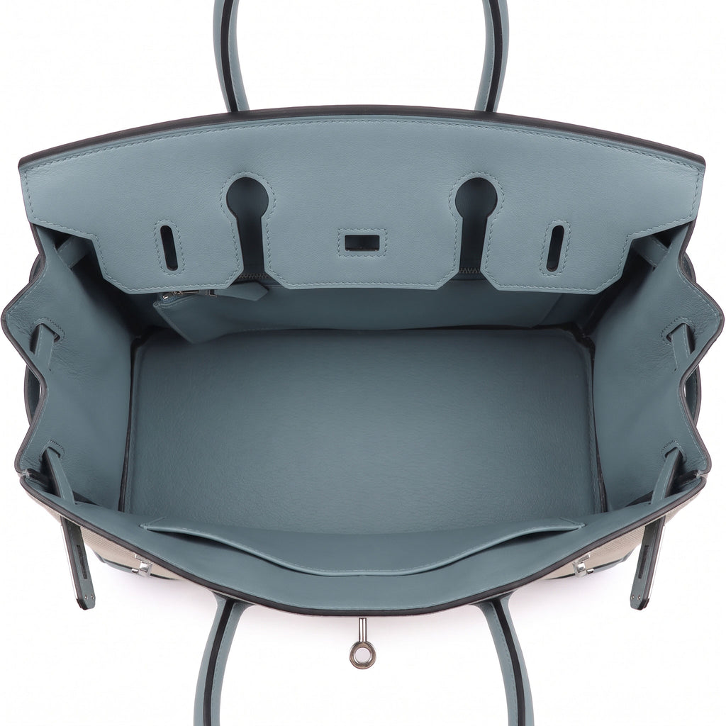 The interior of a structured leather handbag shown from above, featuring a dusty slate blue lining with dark brown or black edging and silver-tone hardware.