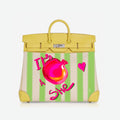 A yellow leather top-handle handbag with a cream and light green striped body, featuring a bright pink pomegranate illustration and the text 'It's She' in pink script on the front.