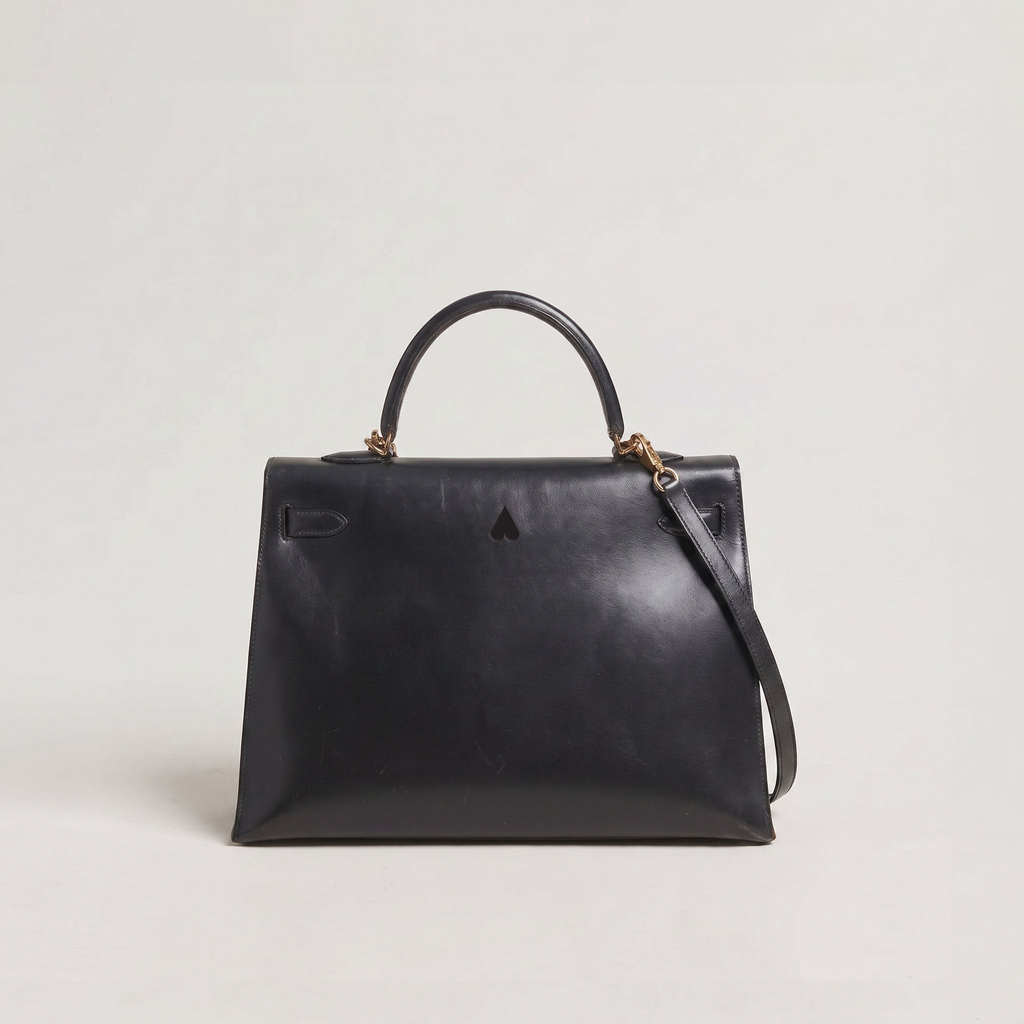 A black leather top-handle handbag with a structured rectangular silhouette, a detachable shoulder strap, and small gold-tone hardware, photographed against a plain light gray studio background.