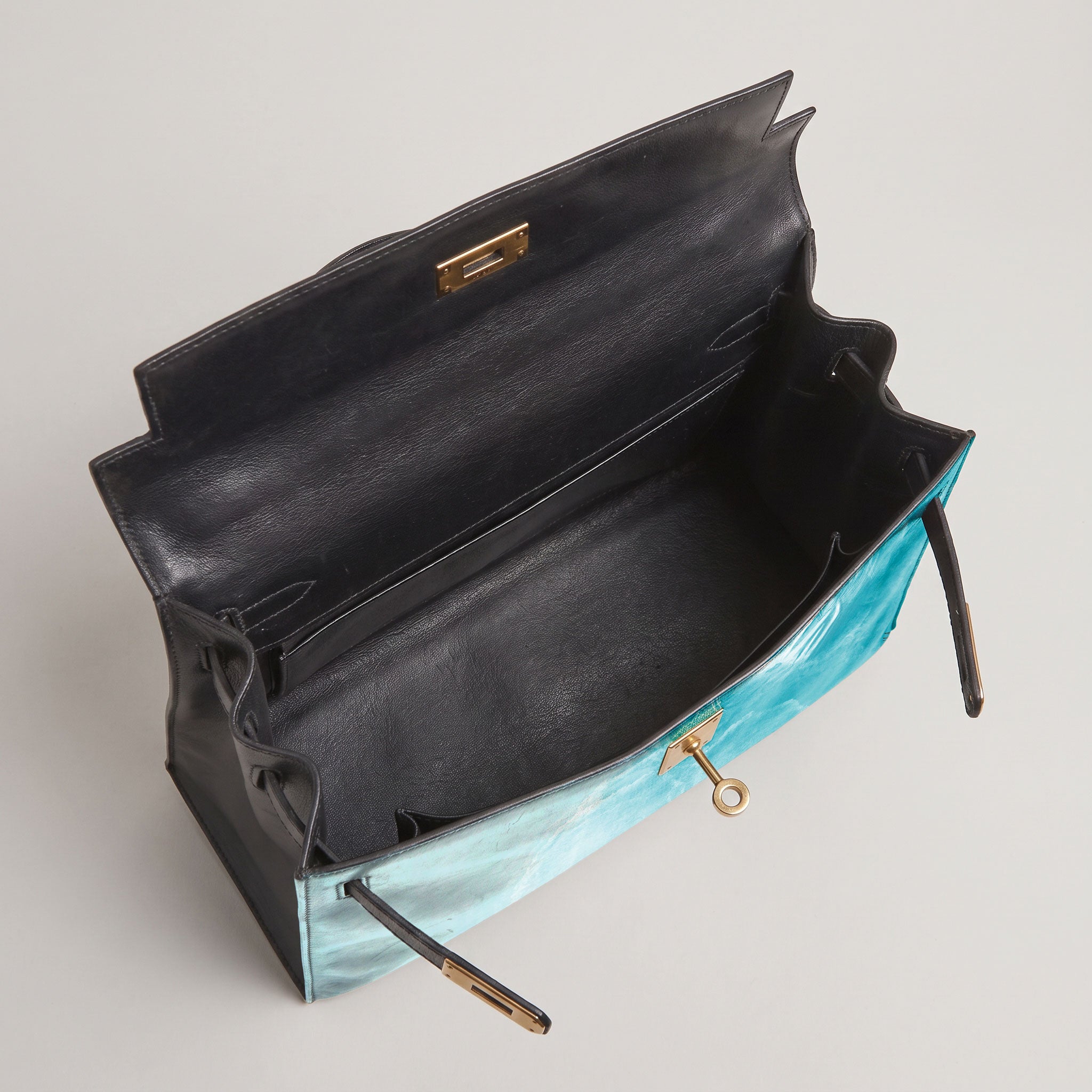 An open luxury handbag with a black leather interior and exterior, accented by a light blue-green marbled leather front panel and gold-tone hardware.