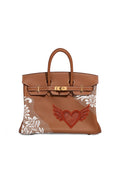 A brown leather handbag with gold-tone hardware, featuring a red embroidered winged heart on the front and white lace applique on the sides, shown against a plain white background.