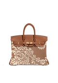 A structured brown leather handbag with an off-white floral lace overlay on the front, featuring gold-tone hardware and dual top handles, shown against a plain white background.