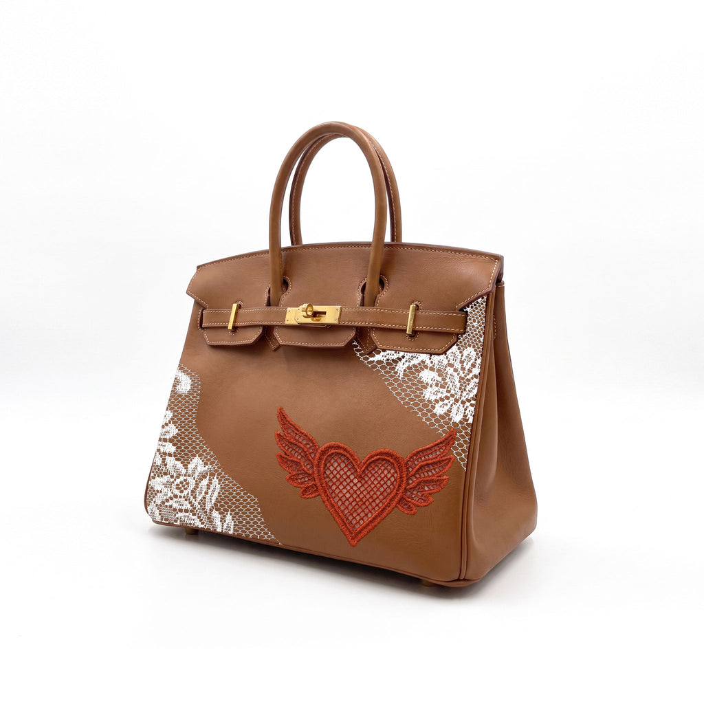 A brown leather handbag with gold-tone hardware, featuring white lace applique on the lower sides and a red embroidered winged heart on the front, set against a plain white background.