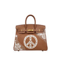 A brown leather handbag with gold-tone hardware, featuring a large white peace sign motif and white floral lace-like patterns on the front, set against a plain white background.