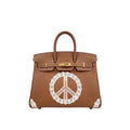 A brown leather handbag with gold-tone hardware and a large white lace peace sign applique on the front, shown against a plain white background.