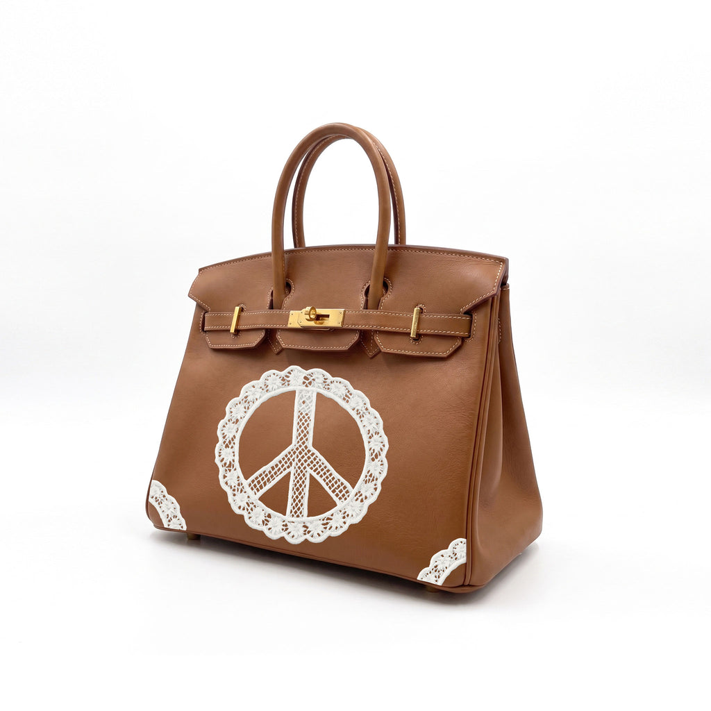 A brown leather handbag with gold-tone hardware and a large white lace-style peace sign applique on the front, photographed on a clean white studio background.