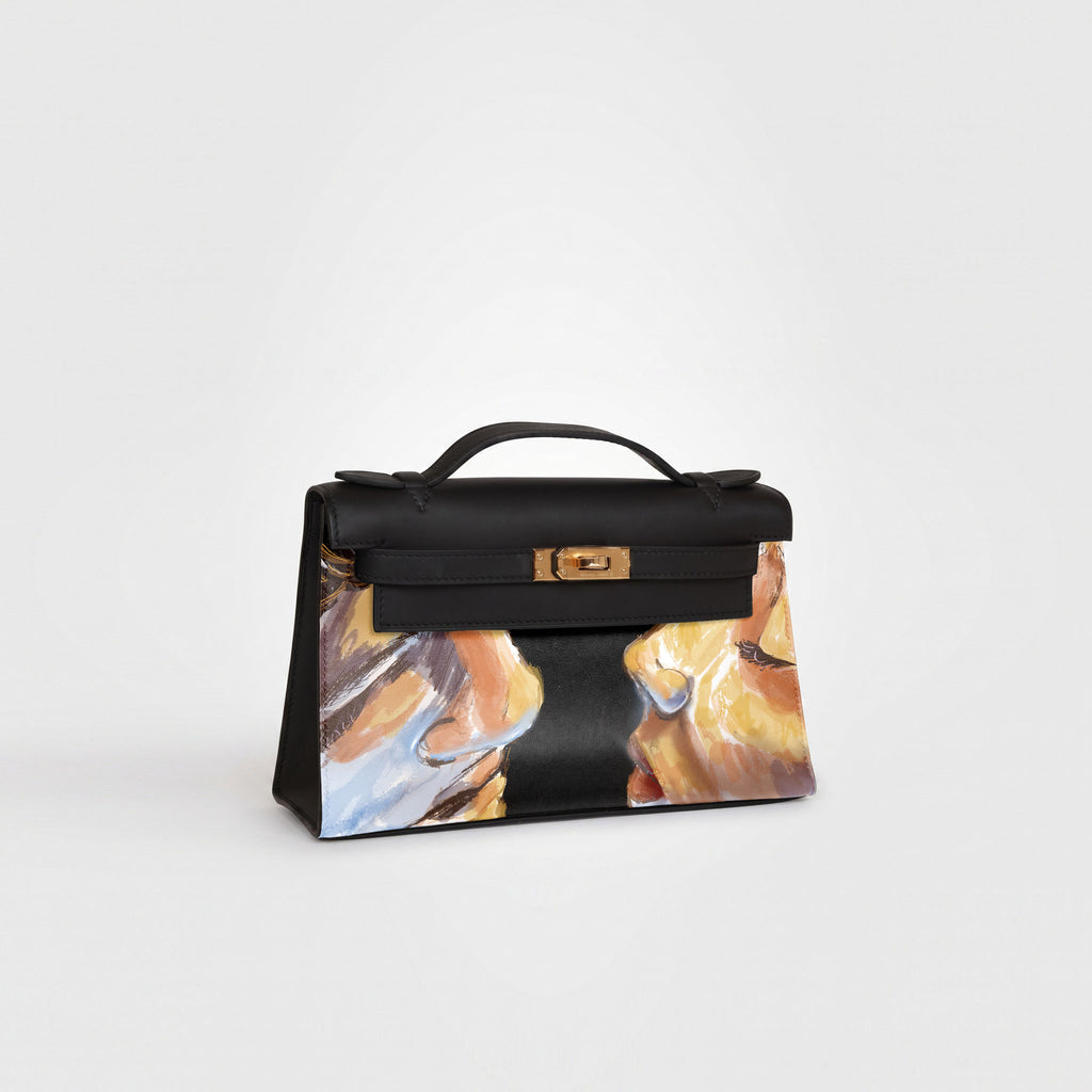 A structured black leather handbag with a gold-tone turn-lock closure and top handle, featuring an abstract painted face design in warm gold, brown, and blue tones on the lower front panel, photographed on a clean light gray background.