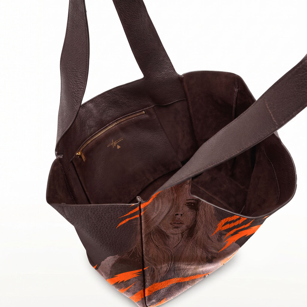 A dark brown leather tote bag with a printed portrait of a woman with long hair and bright orange graphic accents, shown on a clean white background.