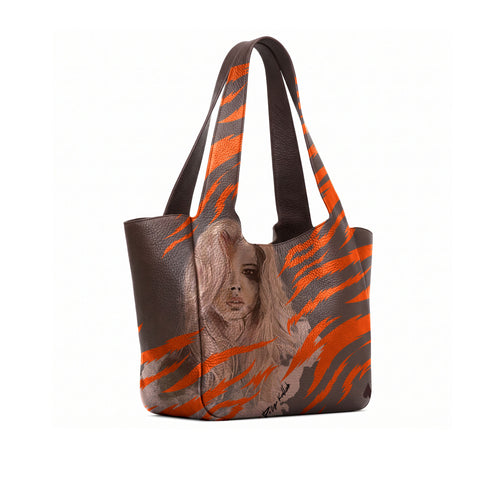 A dark brown leather tote bag featuring a bold orange tiger-stripe pattern and a printed portrait of a woman on the front, set against a plain white background.